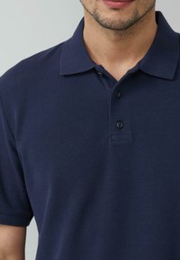 Granatowa koszulka polo z teksturowanej tkaniny, z kołnierzykiem, trzema guzikami i małym zakładanym przodem. Zbliżenie na górną część ciała.