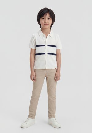 Dječak stoji noseći bijelu košulju kratkih rukava s ovratnikom i tamnoplave pruge, bež hlače i bijele tenisice na jednobojnoj pozadini.