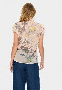 Woman with long blonde hair wearing a light pink floral blouse with puffed short sleeves and dark blue wide-leg pants, facing away.