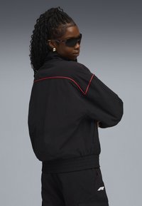 Black windbreaker jacket with red piping, featuring a boxy fit and elastic waistband. Worn with dark sunglasses, showcasing a sleek design.