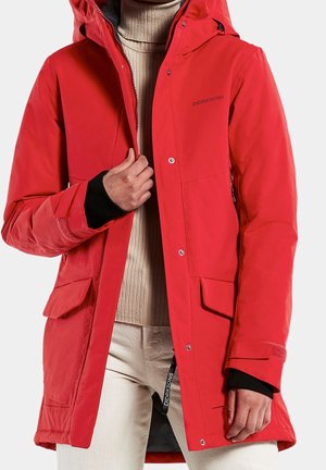 Person trägt einen leuchtend roten Wintermantel mit Kapuze, einen beigen, gerippten Rollkragenpullover und helle Hosen, hält die Vorderseite des Mantels nahe dem Reißverschluss.