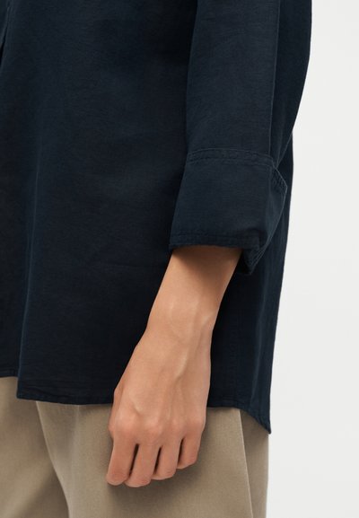 Άτομο που φοράει σκουρόχρωμο μακρυμάνικο πουκάμισο με τα μανίκια τυλιγμένα και μπεζ παντελόνια, το χέρι χαλαρό δίπλα στο σώμα σε φωτεινό φόντο.