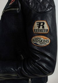 Veste de course en cuir noir avec des détails matelassés aux épaules et deux écussons brodés sur la manche, avec le texte "RACING" et "REDSKINS SERVICE".