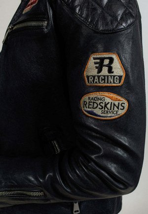 Veste de course en cuir noir avec des détails matelassés aux épaules et deux écussons brodés sur la manche, avec le texte "RACING" et "REDSKINS SERVICE".