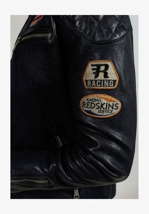 Veste de course en cuir noir avec des détails matelassés aux épaules et deux écussons brodés sur la manche, avec le texte "RACING" et "REDSKINS SERVICE".
