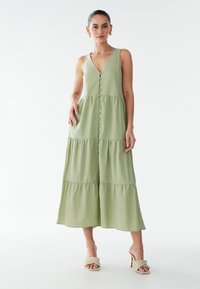 Maxi-jurk van lichtgroffe stof, met een diepe V-hals en knoopsluiting aan de voorkant. Laagjesontwerp met een zachte textuur. Gecombineerd met beige sandalen met hak.