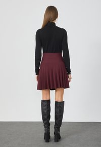 Sort turtleneck bluse, tætsiddende i taljen, parret med en knælang, plisseret bordeaux nederdel. Sorte knæhøje læderstøvler fuldender looket.