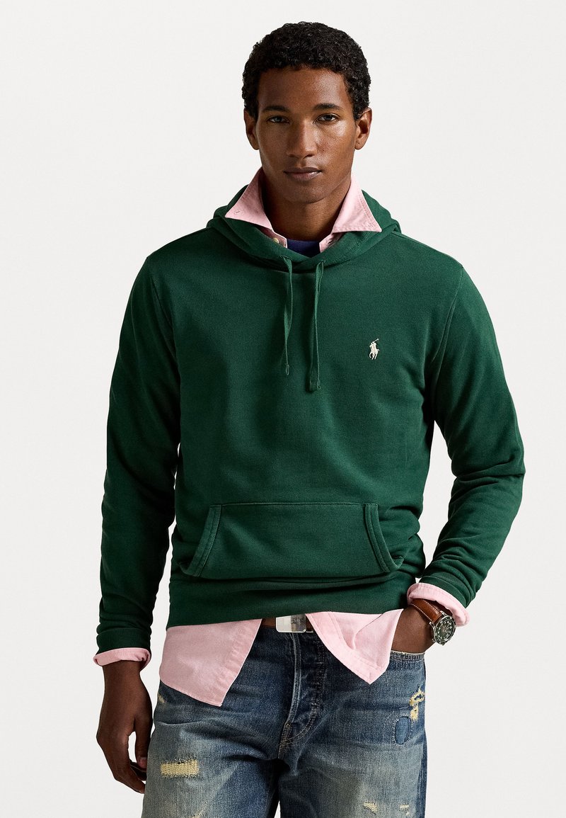 Groene katoenen hoodie met een voorkeep, een capuchon met trekkoord en een wit logo. Gelaagd over een lichtroze overhemd met kraag en gedragen met versleten jeans.