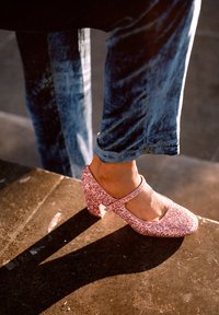 Třpytivé růžové Mary Jane boty s masivním podpatkem a páskem přes nohu, obuté s modrými džínovými kalhotami, opřené o tmavé kamenné schody.