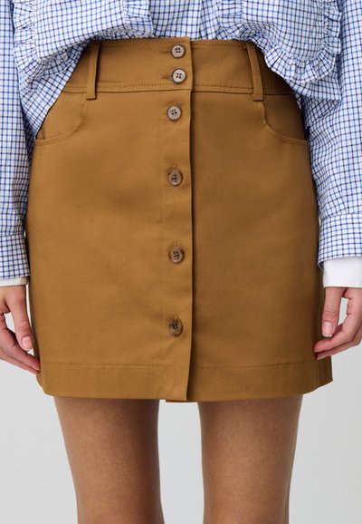 Gonna di cotone marrone con taglio dritto, bottoni frontali e tasche laterali. Abbinata a una camicia a quadri blu e bianca.