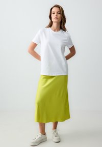 Weißes Baumwoll-T-Shirt mit kurzen Ärmeln, kombiniert mit einem knielangen leuchtend grünen Rock aus einem glatten, fließenden Stoff. Weiße Turnschuhe runden das Outfit ab.