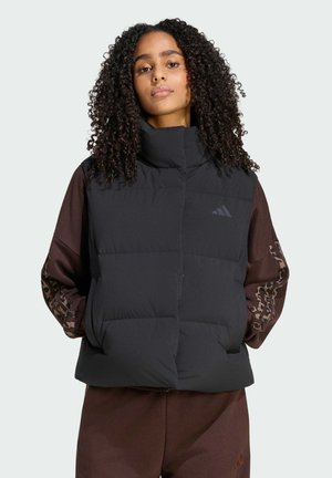 Kvinde med krøllet hår iført en sort quiltet vest over en brun langærmet outfit med mønstrede ærmer, stående mod en ensfarvet baggrund.