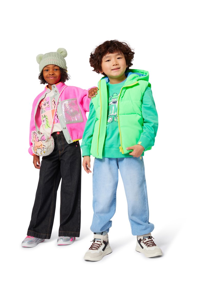To barn står, den ene iført en rosa jakke, grønn lue med pom-pomer, og en enhjørningveske, den andre i en grønn vest og lyseblå jeans.