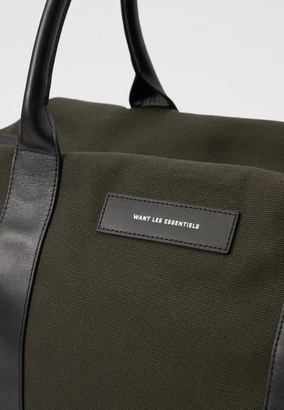 Olivroheline kott, mis on valmistatud tekstuuritud kanvasest koos musta nahast detailidega; sisaldab ristküliku kujulist silti, millel on reljeefne tekst "WANT LES ESSENTIELS."