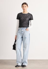 Top transparente con rayas negras superpuesto sobre una camiseta de manga larga blanca, combinado con jeans de pierna ancha desgastados de color azul claro y zapatos de punta negra.