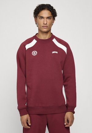 Homme aux cheveux bouclés portant un sweat bordeaux avec des accents blancs aux épaules et le texte "UNFAIR" sur la poitrine sur un fond uni.