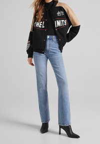 Černá a béžová varsity bunda s vyšitým textem, vzory na rukávech; modré rozšířené džíny a černé boty s špičatou špičkou. Celkově hladké textury.