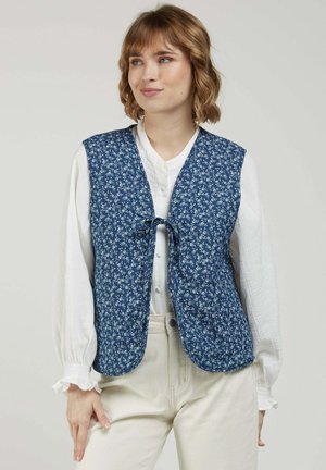 Femme portant un gilet matelassé bleu floral par-dessus une blouse blanche à manches longues avec des poignets volantés et un pantalon de couleur claire, debout devant un fond uni.