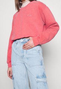 Osoba v korálové mikině a světle modrých denimech s cargo kapsami, s jednou rukou opřenou o bok a viditelným bílým volánkovým límcem.