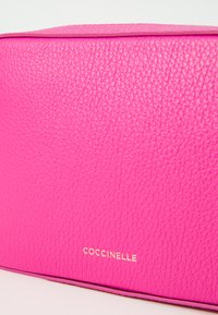 Coccinelle pink leather bag with textured surface, rectangular shape, and gold logo on the front. Features smooth edges and clean lines.
