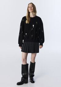 Czarny sweatshirt z kluczowymi motywami, zestawiony z plisowaną szaryą spódnicą i czarnymi skórzanymi kozakami do kolan. Gładka tekstura, luźny krój.