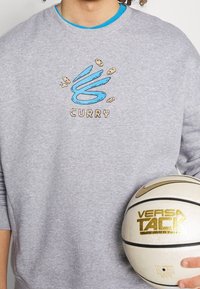 Sweatshirt gris avec col rond, arborant un design éclaboussé en bleu brodé avec "CURRY" en lettres tan. Tenant un ballon de basket beige et or.