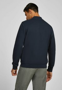 Marineblaues Sweatshirt mit Rundhalsausschnitt und gerippten Bündchen. Texturiertes Material, lässige Passform, sichtbares Logo auf der Rückseite. Graue gemusterte Hose.