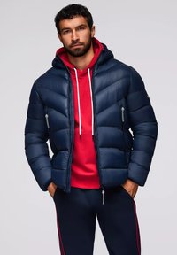 Giacca imbottita blu navy con un design strutturato, caratterizzata da sezioni trapuntate orizzontali, chiusura con zip e tasche laterali. Felpa con cappuccio rossa sotto.