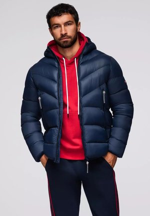 Marinblå pufferjakke med et struktureret design, der har horisontale quiltede sektioner, lynlås lukning og sidelommer. Rød hættetrøje under.