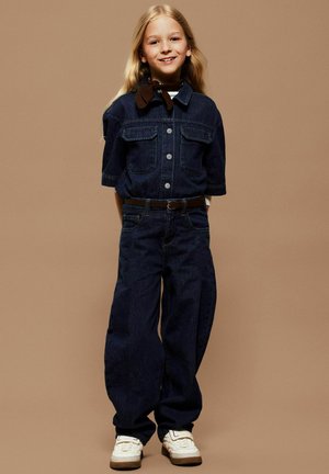 Barn med langt blondt hår som smiler, iført mørk denimskjorte og bukser, brun skjerf, hvite joggesko, står foran en beige bakgrunn.