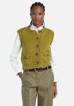 Knit olive green vest with a round neck, button front, and two front pockets. Worn over a white shirt and checked trousers, featuring a textured finish.