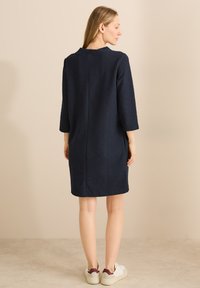 Nachtblaues, strukturiertes Kleid mit drei Viertelärmeln, geradem Schnitt, minimalistischem Rückennähten, kombiniert mit weißen Turnschuhen.