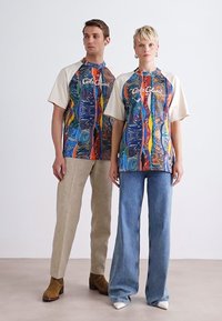 Farverige, abstrakte mønstrede oversized T-shirts med beige ærmer; modeleret med lyse bukser og brune støvler. Bemærkelsesværdig grafisk logo viser sig.