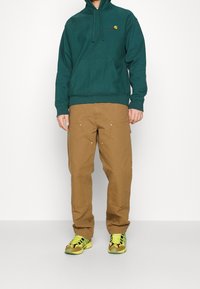 Grøn bomulds hoodie med frontlomme, brune cargo bukser med lommer og metal accenter, samt multikolore sneakers.