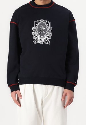 Homme portant un sweat-shirt noir avec un emblème blanc et le texte "Hilfiger", associé à un pantalon blanc, debout devant un fond uni.