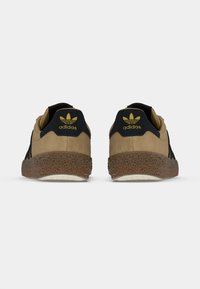 Καφέ σουέντ αθλητικά παπούτσια με μαύρες λεπτομέρειες, που διαθέτουν υφασμένο, σχεδιασμένο καουτσούκ στη σόλα και χρυσό λογότυπο adidas στη φτέρνα.