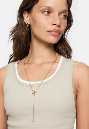 Gold necklace with a unique pendant design. The model wears a light beige tank top with a white accent on the neckline, showcasing the jewelry.