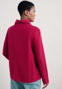 Pullover fucsia con colletto alto, maniche lunghe e tessuto texturizzato. L'orlo è leggermente curvato sul retro.