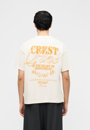 Άτομο με κοντά μαύρα μαλλιά φοράει λευκό μπλουζάκι με πορτοκαλί κείμενο που γράφει "CREST Pine στην καρδιά του Moosehead" στην πλάτη.