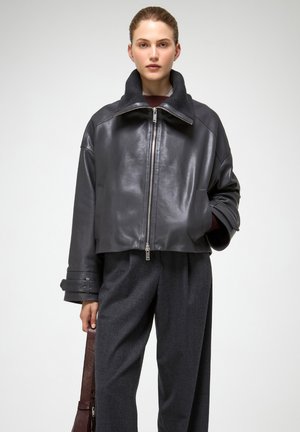 Giubbotto corto in pelle nera con colletto alto, chiusura con zip e maniche ampie. Abbinato a pantaloni in lana grigia e una borsa in pelle marrone.