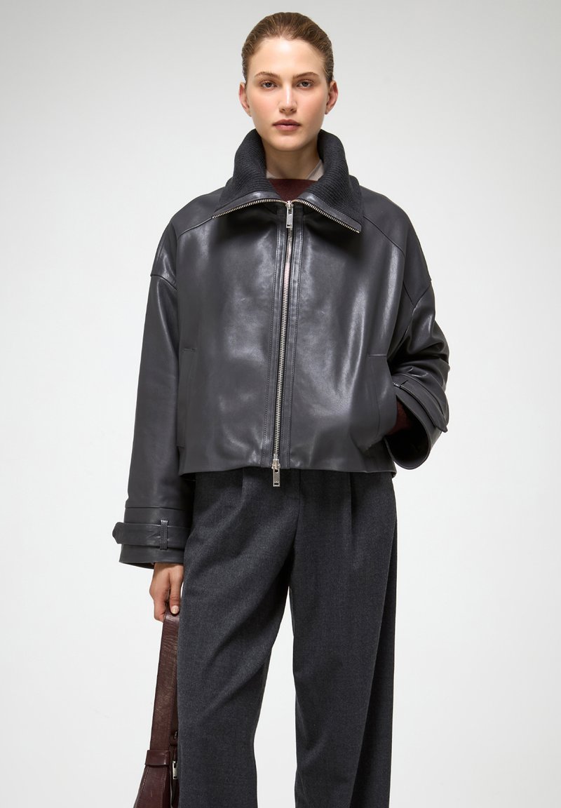 Giubbotto corto in pelle nera con colletto alto, chiusura con zip e maniche ampie. Abbinato a pantaloni in lana grigia e una borsa in pelle marrone.