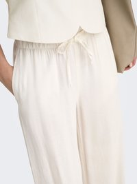 Gros plan d'une personne portant un pantalon blanc ample taille haute avec une taille à cordon et une veste courte crème.