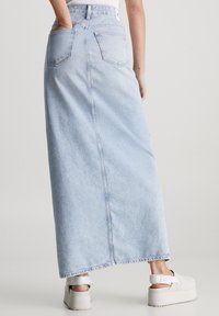 Falda maxi de mezclilla azul claro con dos bolsillos traseros, una costura central en la parte posterior y un dobladillo sin terminar; combinada con sandalias de plataforma blancas.