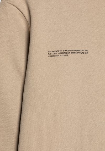 Sudadera de algodón orgánico beige con una textura suave. Presenta un pequeño detalle en negro con texto sobre el tratamiento de la tela para mantener la frescura.