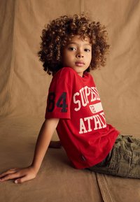 Rød bomulds t-shirt med "SUPREME ATHLETIC" print, sorte detaljer og nummer "84" på ærmet; parret med olivengrønne cargoshorts.