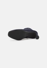 Zapato de ante azul marino con puntera puntiaguda, tacón bajo apilado y suela de goma. Presenta una textura suave con hardware y adornos mínimos.