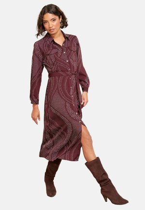 Burgunderfarbenes gemustertes Kleid mit langen Ärmeln, Knopfleiste und Taillengürtel; verfügt über einen hohen Schlitz und eine subtile Textur. Trägt braune kniehohe Stiefel.