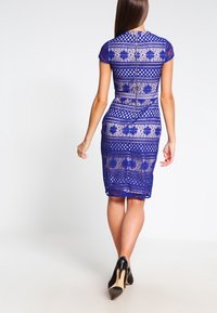 Blue lace dress featuring short sleeves, intricate patterns, and a back zipper. The fitted design extends to knee-length, paired with black heels.