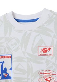 Wit T-shirt met korte mouwen en een grijs geometrisch patroon, uitgerust met rode Superman-logo's en een blauwe kraagaccent. Zacht katoen materiaal.