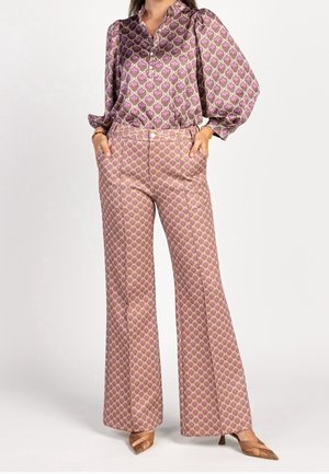 Nainen, jolla on päällään yhteen sopiva vaaleanpunainen ja beige kuvioitu paita, jossa on liehukkeet hihansuissa, ja korkeavyötäröiset leveälahkeiset housut, seisomassa käsien ollessa taskuissa.